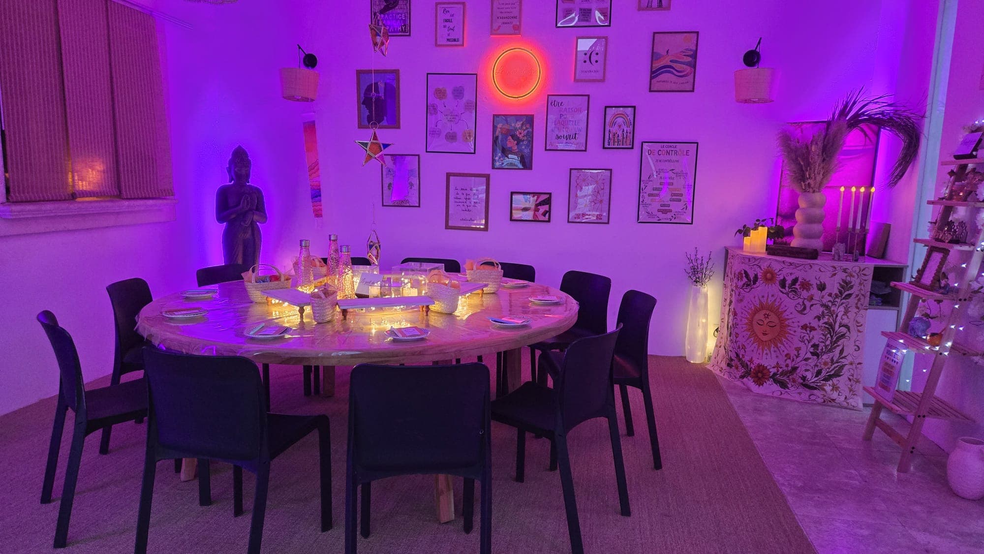 Vue d'ensemble de la salle : grande table ronde dressée, chaises, mur de cadres et desserte, dans une lumière violette